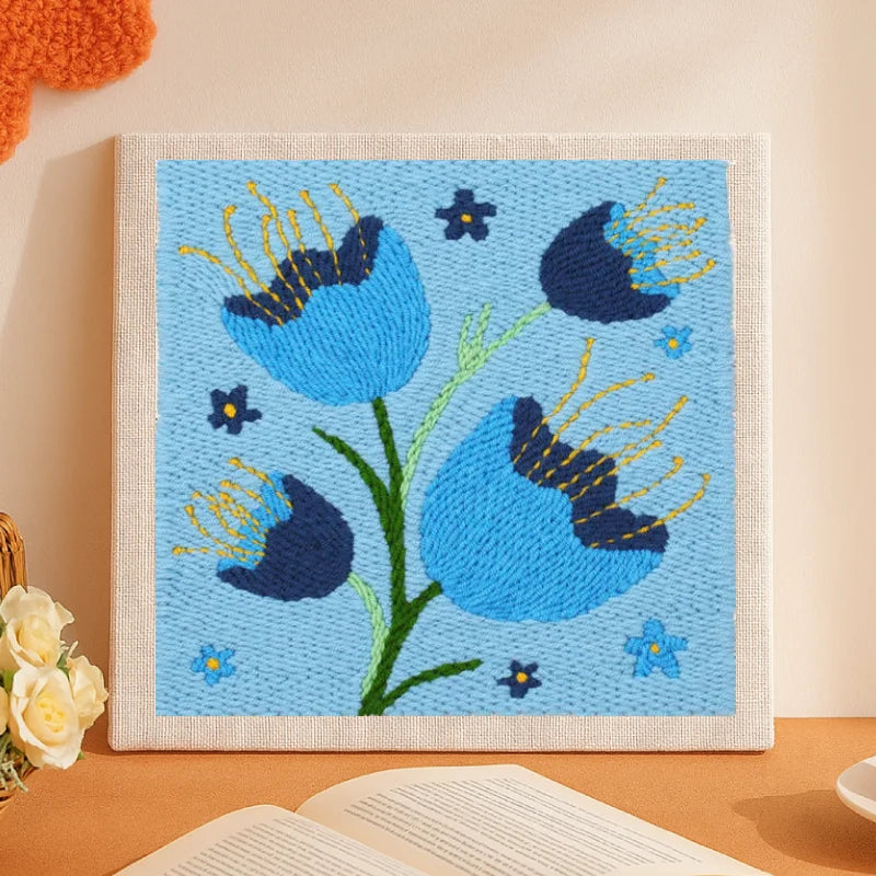 This punch needle artwork showcases vibrant blue flowers with dark navy accents, standing tall on fresh green stems against a light blue background. The yellow embroidery details add a lively sparkle, giving the design a calm yet uplifting charm — perfect for adding serenity to any room.