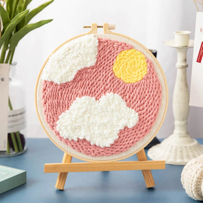 Woven circular art piece with pink background, white clouds, and yellow sun on a wooden stand.