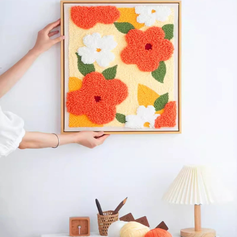 Framed floral artwork with red, white, and green flowers on a yellow background, held by a person against a white wall.