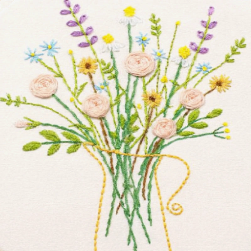 This close-up image highlights the delicate embroidery details of a floral bouquet. Soft pink roses are stitched with spiral techniques, giving them a subtle, textured look. Light blue daisies, tiny yellow blossoms, and lavender sprigs add gentle colour accents, creating a fresh and natural palette. The green stems flow gracefully, while the simple outline of a yellow-threaded jug adds balance and warmth. Together, the composition feels light, calm, and beautifully pastoral — a perfect reflection of handmad