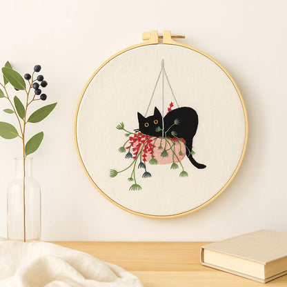 Embroidery hoop with a black cat design hanging from plants on a light background