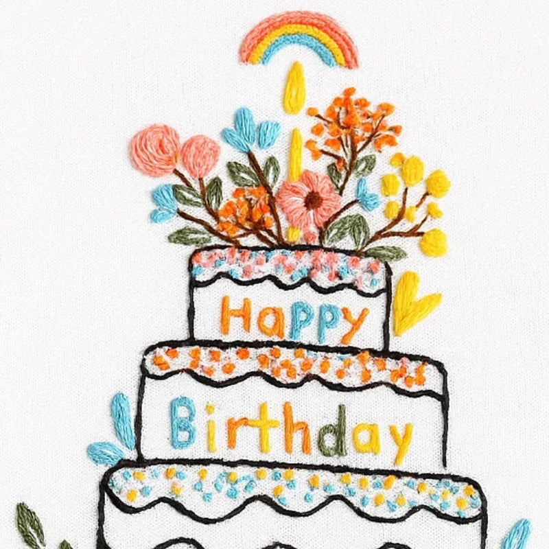 Embroidered design of a birthday cake with flowers, candles, and a rainbow on a white background.