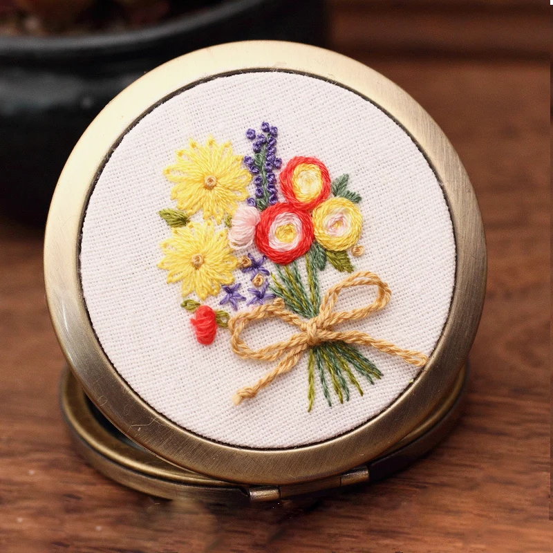 This hand-embroidered compact mirror features a radiant bouquet of yellow daisies, red ranunculus, and touches of lavender — tied together with a soft golden bow. The vibrant colors bring a sense of sunshine and warmth, making it a perfect accessory to brighten your day or to gift as a token of cheer.