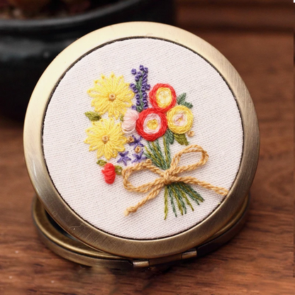 This hand-embroidered compact mirror features a radiant bouquet of yellow daisies, red ranunculus, and touches of lavender — tied together with a soft golden bow. The vibrant colors bring a sense of sunshine and warmth, making it a perfect accessory to brighten your day or to gift as a token of cheer.