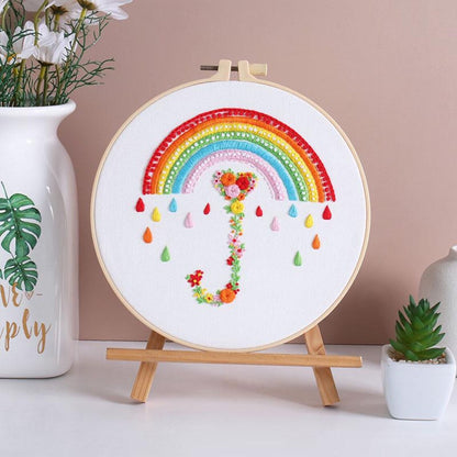 Embroidery hoop with a colorful rainbow and floral design on a wooden stand against a beige wall.