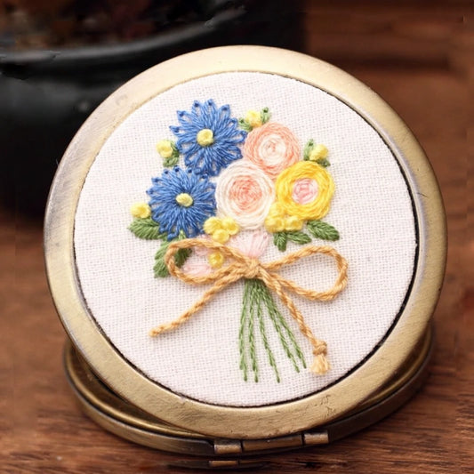 This lovely hand-embroidered compact mirror features a bouquet of blue daisies, yellow blossoms, and soft peach roses tied with a gentle golden bow. The design captures the freshness of spring mornings — vibrant yet soothing. Perfectly sized to slip into your purse, it brings a touch of handmade charm and floral serenity wherever you go.