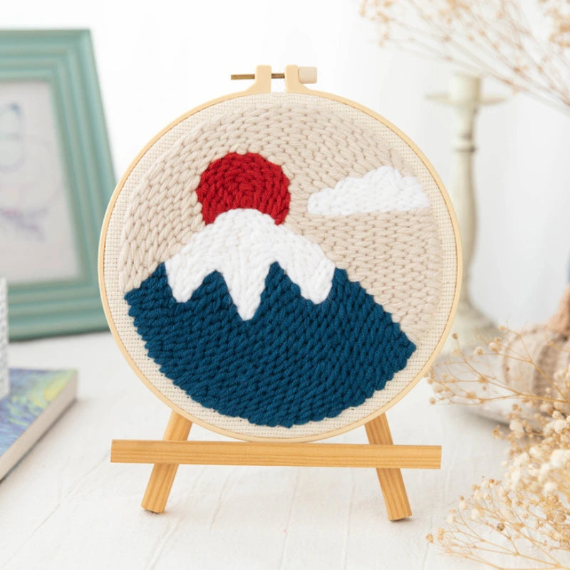 Handmade needlepoint of a mountain scene with a red peak, white snow, and blue mountains on a wooden stand.