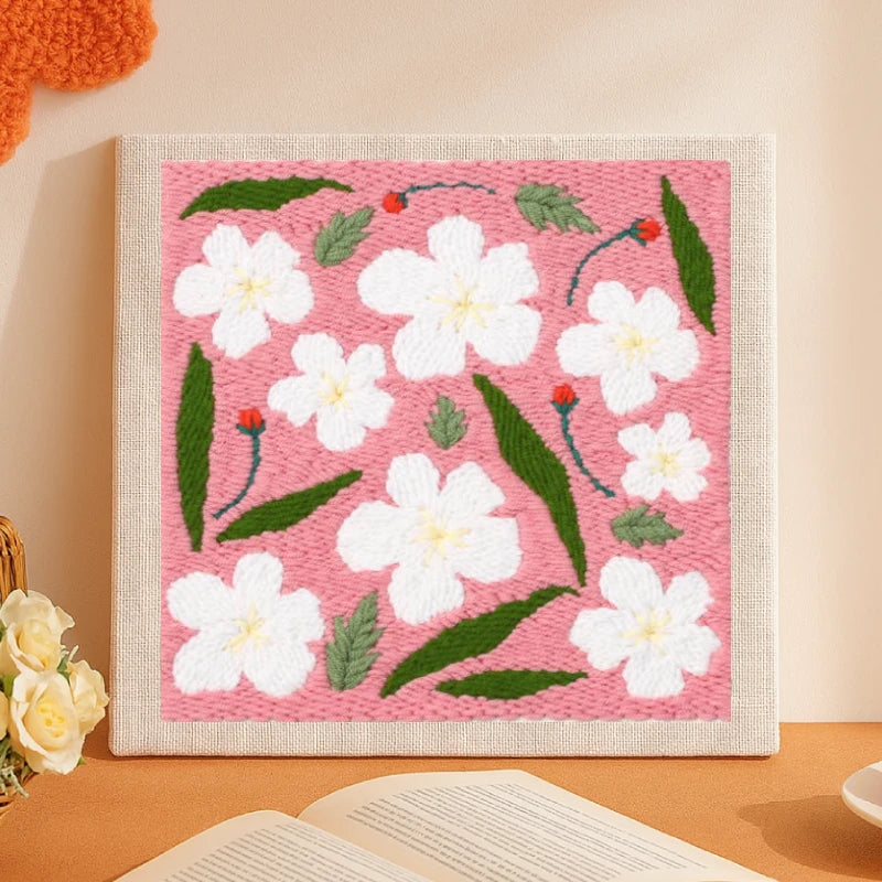 This delightful punch needle piece features white blossoms scattered across a soft pink background, paired with green leaves and tiny red buds.