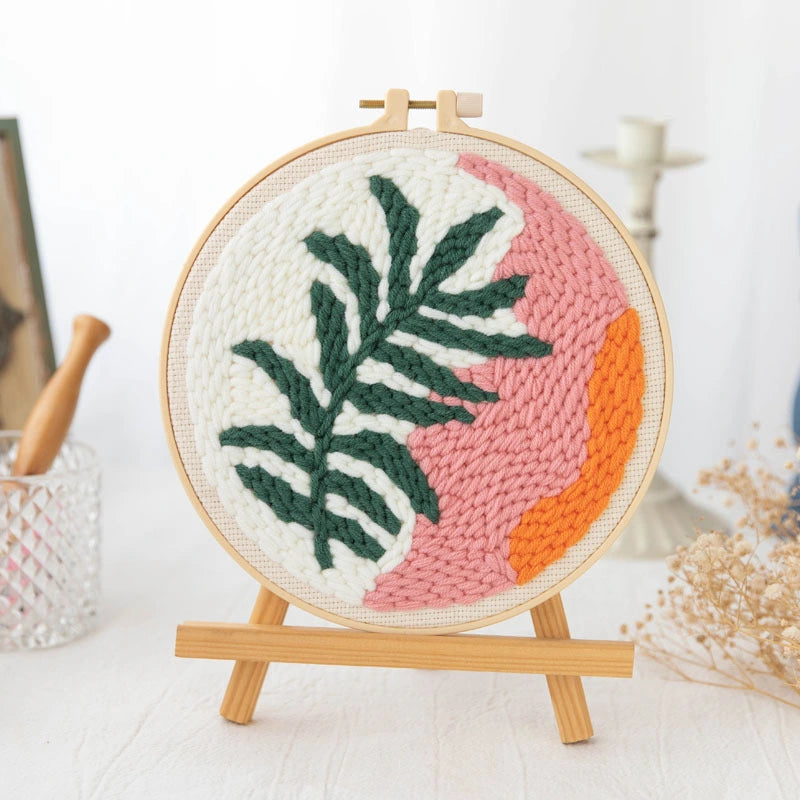 Woven circular art piece with green leaf design and pink and orange abstract patterns on a wooden stand.