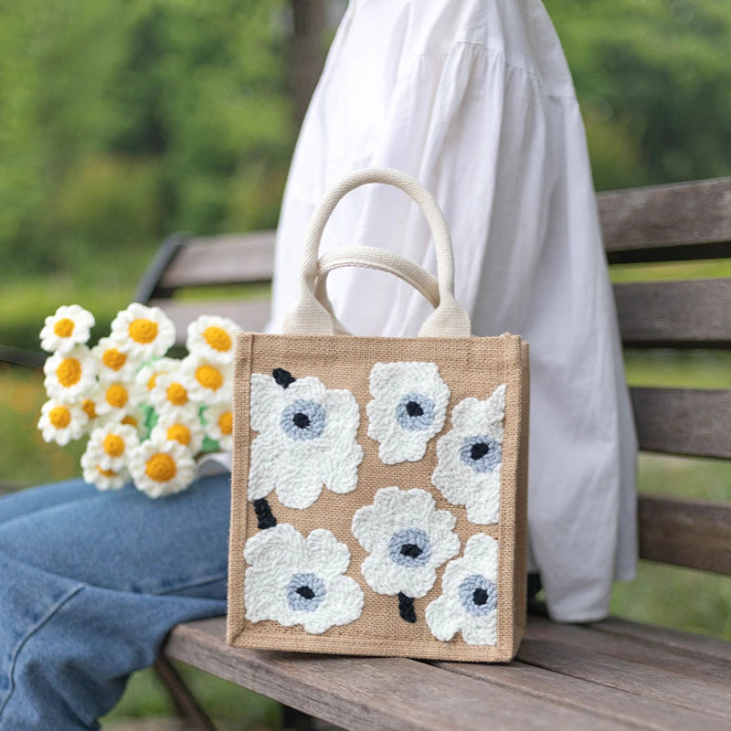 Jute bag with floral embroidery held by a person sitting on a bench outdoors.
